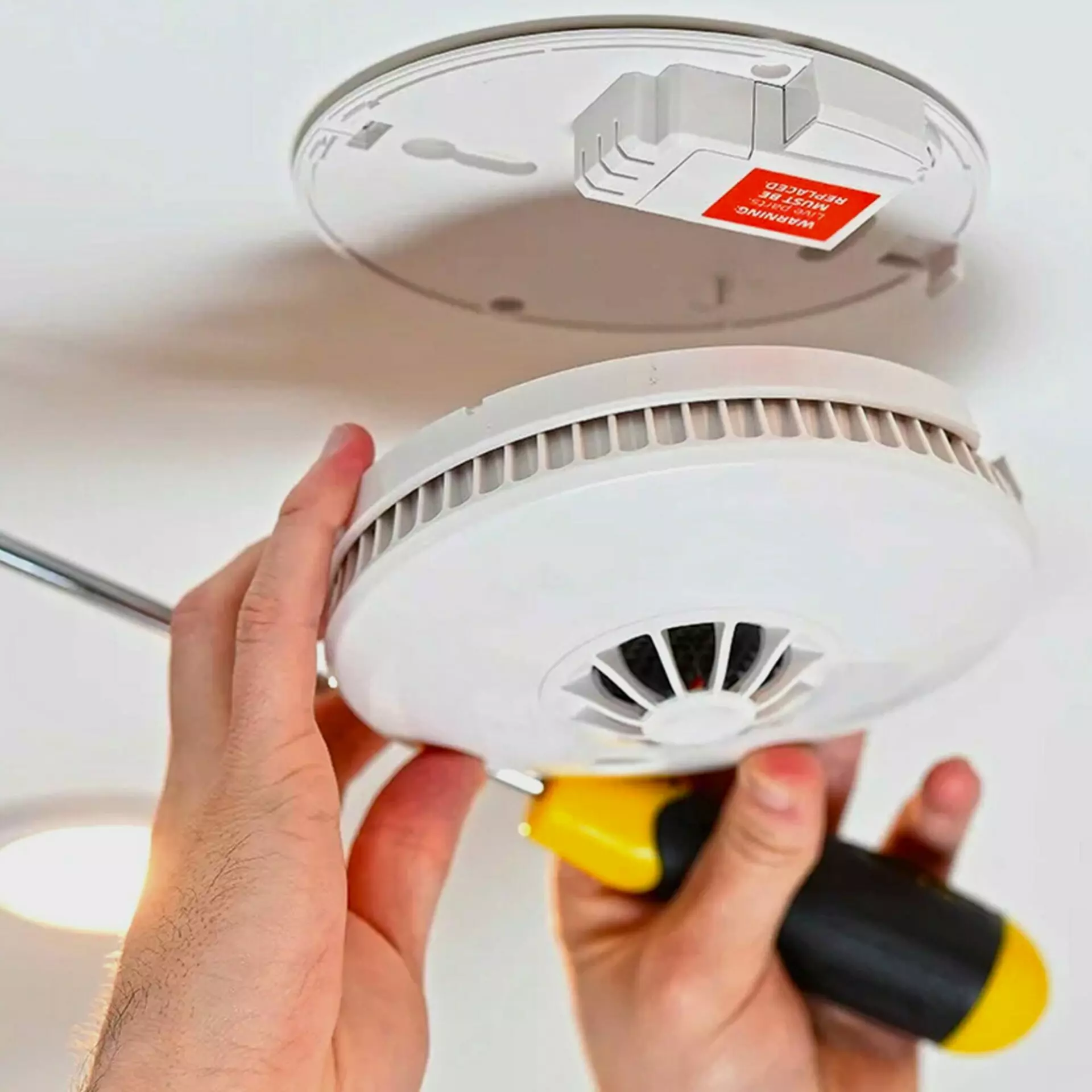 Image has a smoke detector, screwdriver and a person fitting it