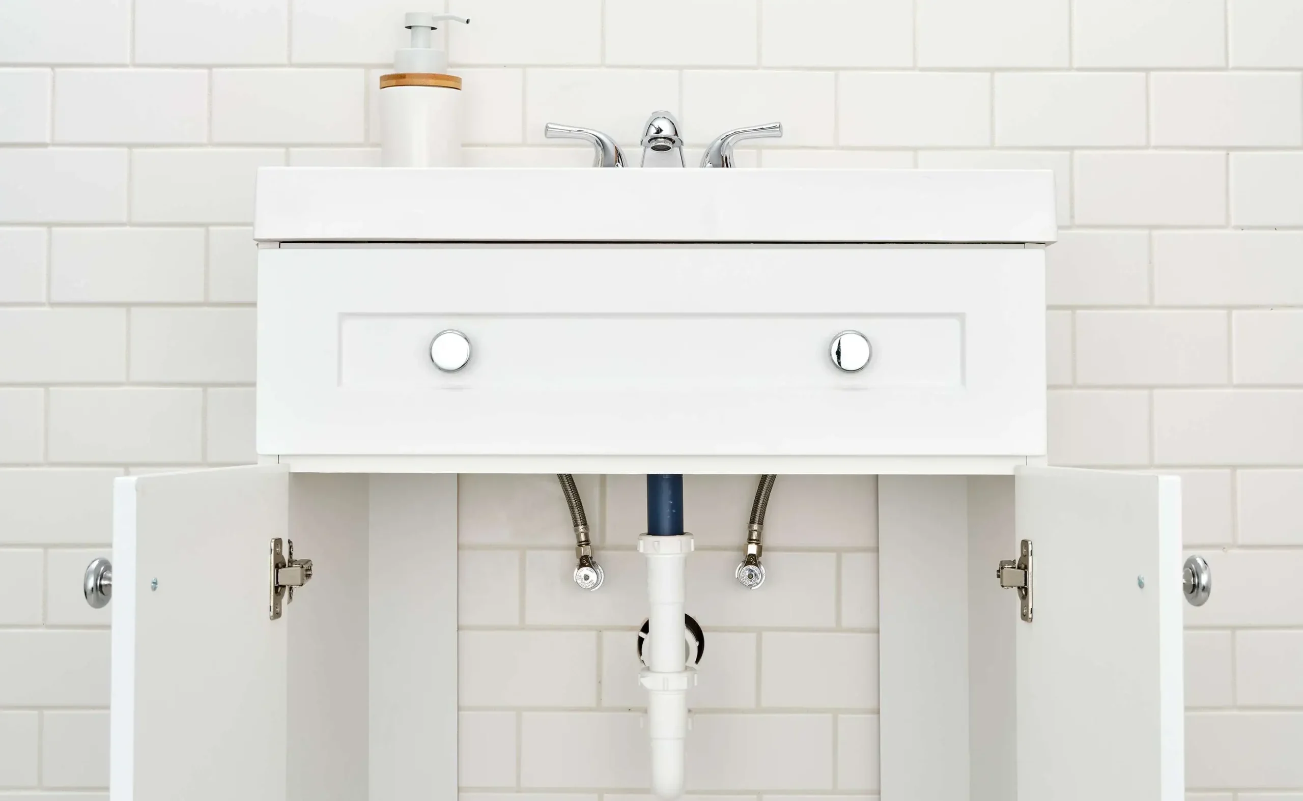 TJ Handyman Services bathroom vanity installation showing a white sink cabinet with open doors, exposed plumbing connections, and a chrome faucet.